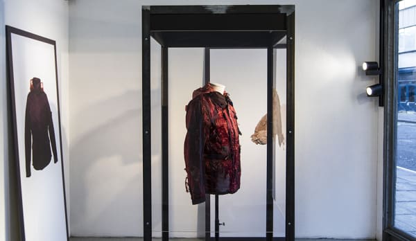 a red and black jacket suspended in a display case. To the right a window revealing a street. To the left (behind the display case) a framed photography of the same jacket.