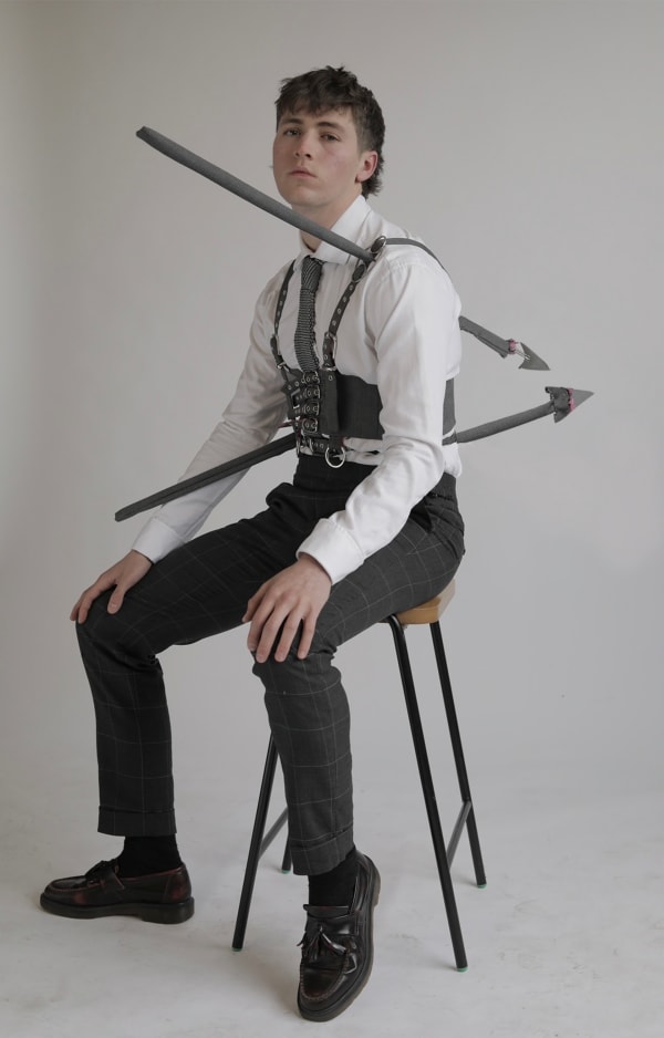 A model sat on a chair wearing a harness with arrows penetrating their body a multi-coloured sculpture