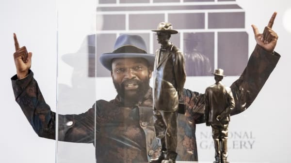 A Black man with his hands up with fingers pointing upwards in front of two bronze-coloured statues of men in hats. 
