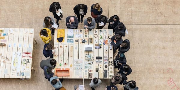 Birds eye view of 2 tutors and a ring of students around a table discussing various objects