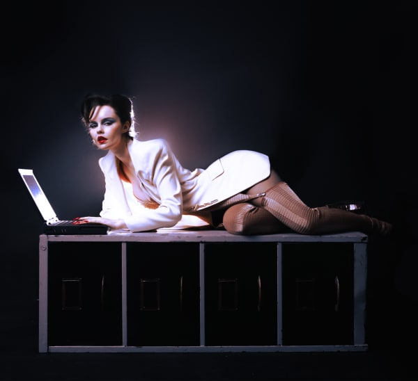 Woman in white suit is lying across a filing cabinet posing and typing on a vintage laptop 