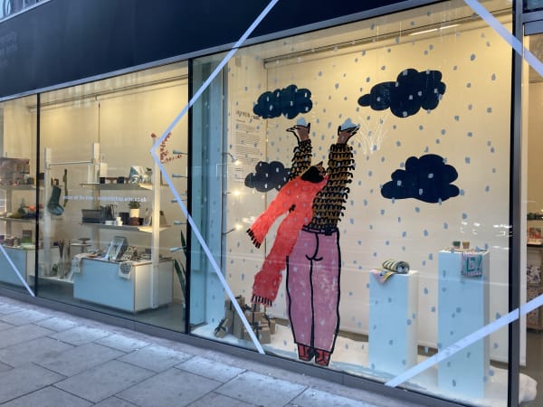 A photo of a window display where a person in a red jumper reaches up to catch falling snow