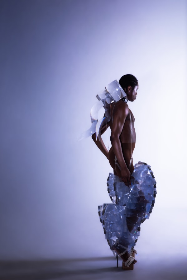 An editorial shot of a Black male model wearing a sculptural transparent garment against a purple lit background