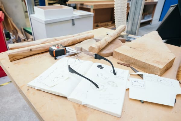 A close up of a wooden table with materials and implements for working with wood, plus a sketchbook with some illustrations and diagrams