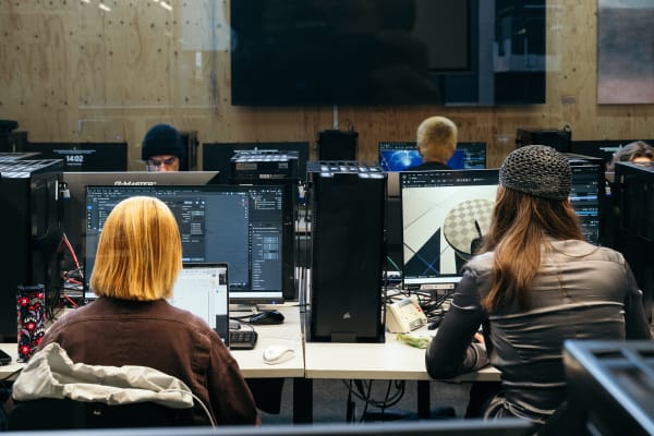 A student working on a desktop computer in a partitioned booth. They are wearing a black baseball cap and  there is a fashion project on screen. To their right is an upright tablet charging with a black screen.