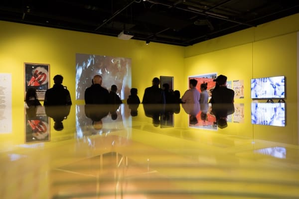 Photo of a group of people looking at a collection of paintings displayed on a yellow wall