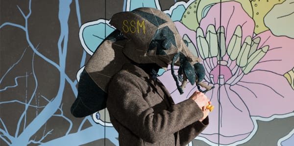 Photograph of a man wearing a giant fly mask in front of a flower mural