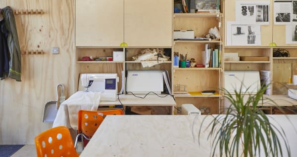 A informal but well organised workshop with plywood walls and cupboards, orange chairs, a sewing machine and a tabletop in the foreground