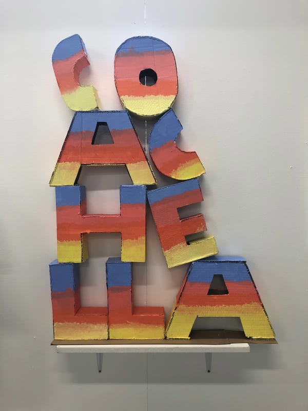 cut out letters painted with red, yellow, pink and orange horizontal stripes. They spell out 'Coachella' and are stacked in a triangular formation 