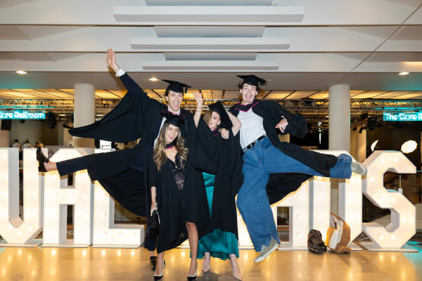 Four students posing in front of a lightbox that reads 'UAL Grads'