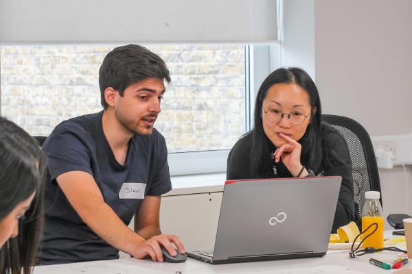 2 people sit at a table talking and looking at a computer. They are talking and looking intrigued. 