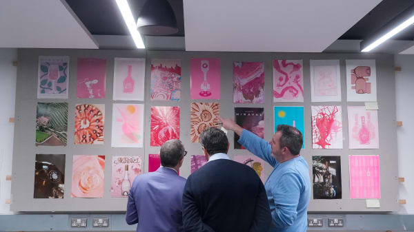Three men looking at artworks created in mostly pink colours on a wall 