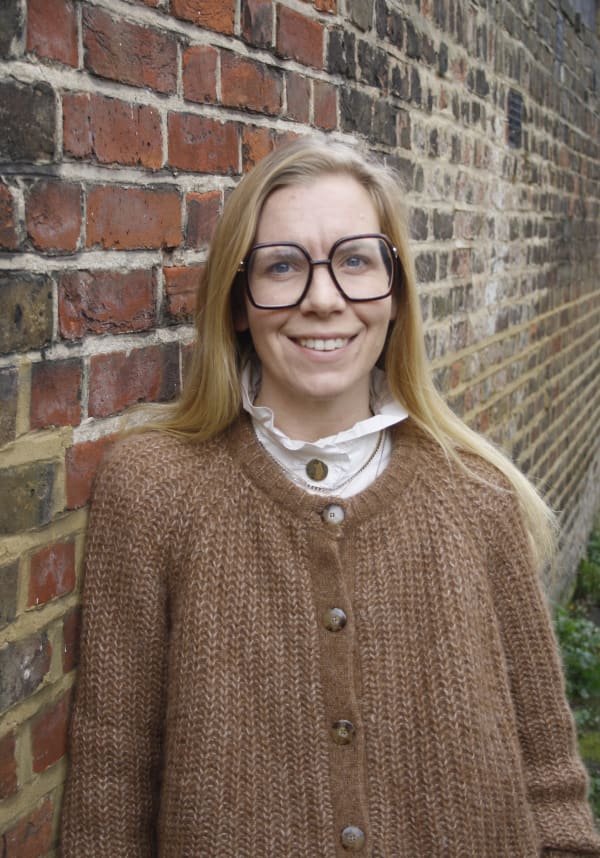 Francesca Shepherd stands in front of a brick wall. She has blond hair, wears large dark rimmed glasses, a white shirt and camel coloured cardigan.
