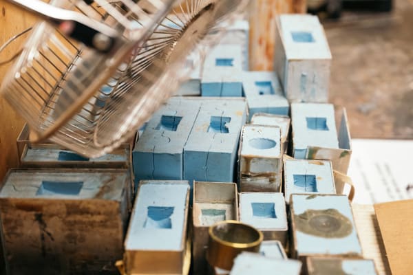 A collection of small, rectangular metal casting moulds arranged closely together on a work surface. Each mould has a blue interior cavity, with different shapes and cutoutsused to form specific parts. Above is a metal wire fan or protective grille, partially in view, used for cooling and ventilation. In the shot we can see signs of residue from casting materials.