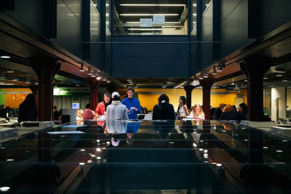 A group of students are perched together in the library and behind them, others are seated in rows of desks. To the forefront of the image is a large reflective table surface 