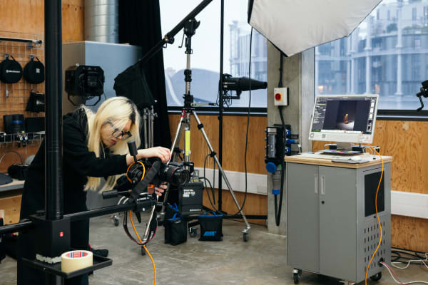 A student is setting up a camera on a tripod in the studio. There is a pedestal with a screen to the right in front of 2 windows. Behind her there are hanging accessories and equipment. 