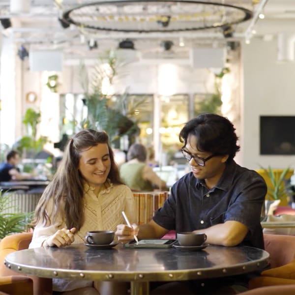 A man and a woman sit at a table collaborating and drawing on a tablet. Others work in the background.