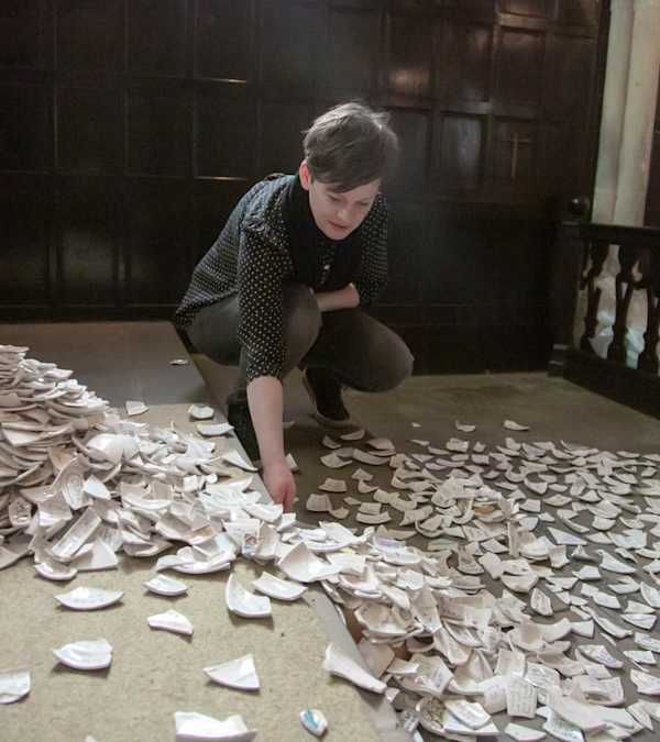 Sarah is kneeling in front of a huge pile of small irregular ceramic pieces with text on them inside a cathedral setting 