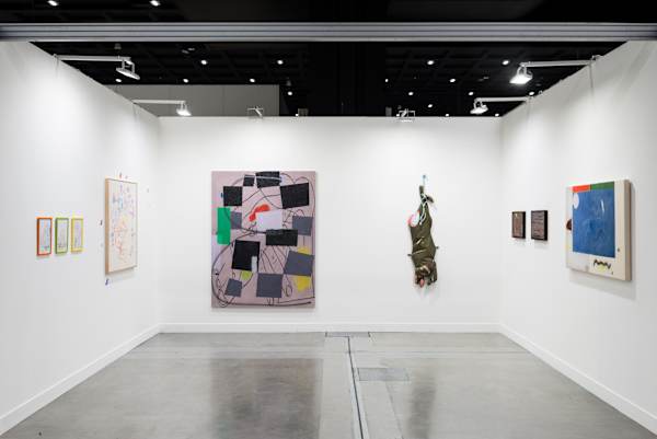 Installation view of an art fair booth featuring abstract paintings, framed works and a hanging sculptural piece displayed on white walls under gallery lighting.