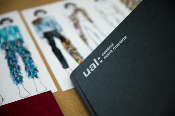 Black notebook with UAL Central Saint Martins Logo. Students work in background