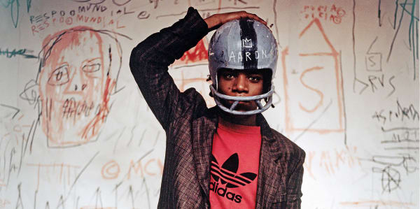 Photo of Jean-Michel Basquiat standing in front of a white wall with drawings on, wearing a helmet with one hand on his head