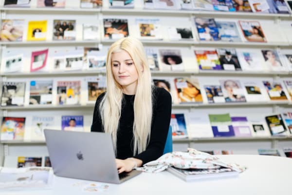 A young blonde woman sits at a desk working on her laptop