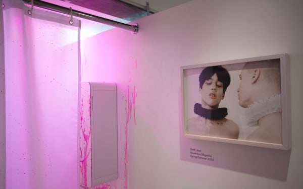 exhibition view of a photo of two topless models wearing ruff collars in black and white. In the background a pink ink stained wall.