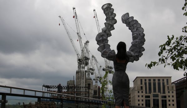 A model wearing sculptural armbands facing cranes against a London skyline