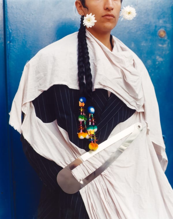 person in a suit with cloth draped over and pinned with a giant hair clip against a bright blue backfrop