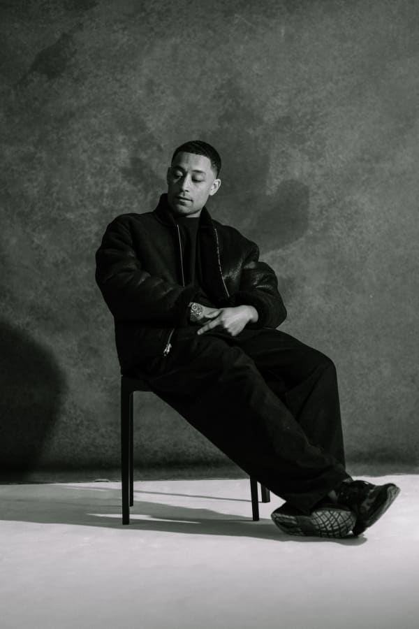 Black and white photograph Loyle Carner. Loyle sits in a chair look down and away from the camera. He is short hair and wears dark clothing and shoes.