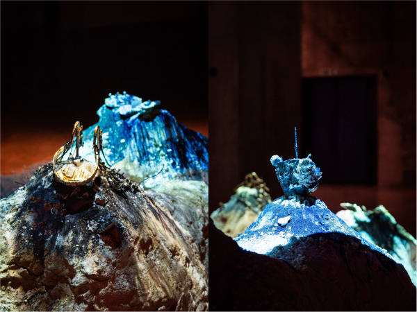 Spotlit installation shot of artworks by Christina Peake. Small metal and clay sculptures top mounds of glittering blue sand.
