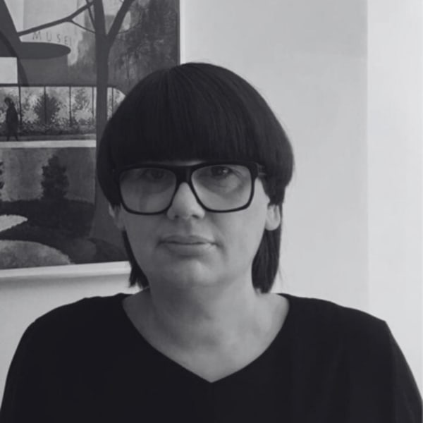 Sally Denton, looking at the camera wearing thick black rimmed glasses, standing in front of a wall with a painting on it. 