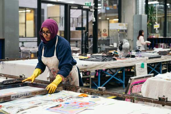 A student in a headscarf and heavily dye spattered apron is holding a wooden frame with a floral print design inside it on a long table with more printworks and pots of coloured dye and a scissors. In the background other students are working on their own projects and we can see industrial flooring with yellow markings.. 
