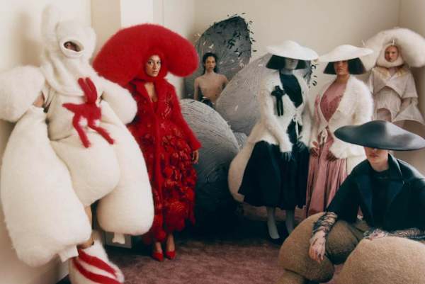 A group of models pose in an indoor setting wearing elaborate, sculptural avant-garde outfits and oversized hats.