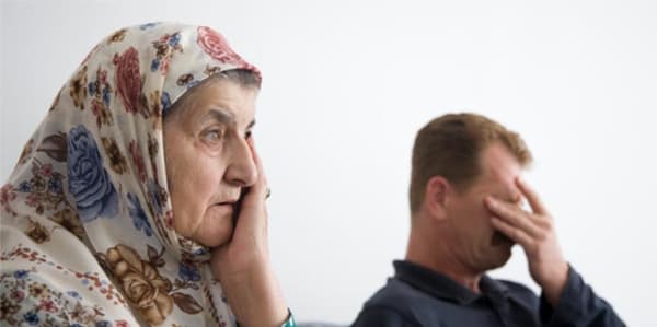 A photograph of 2 people, an older lady in the foreground resting her palm on her face. In the background a man that appears to be rubbing his eyes with his hand, and frowning as though he is wiping tears.