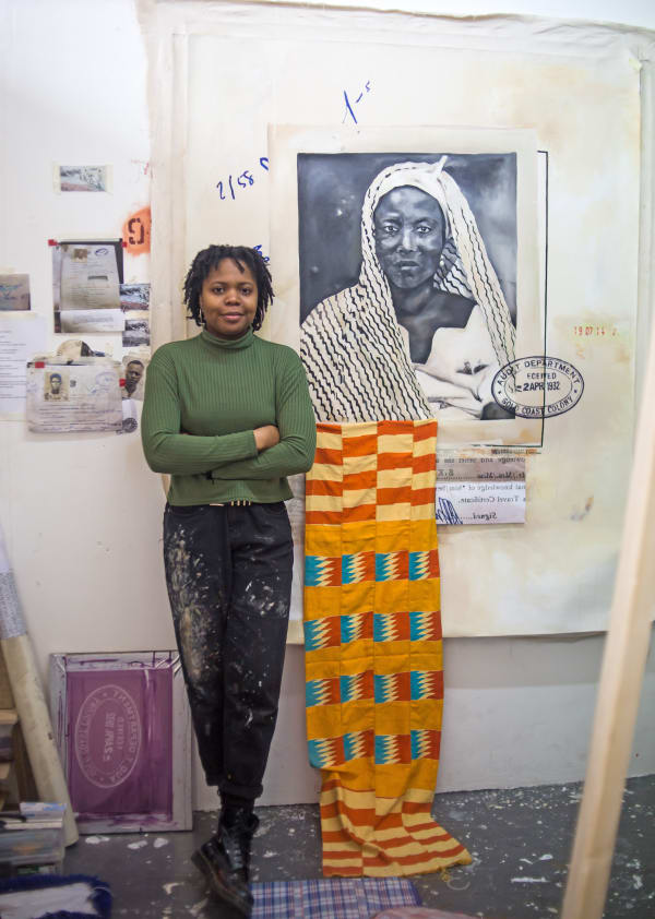 Woman leaning against a white wall in a studio, next to black and white painted portrait of a woman and bright yellow and orange patterned fabric draped down to the floor.