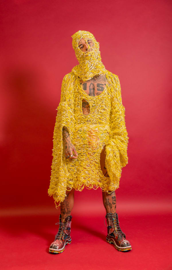 Male model wearing yellow knitted tunic with face mask in front of a pink background