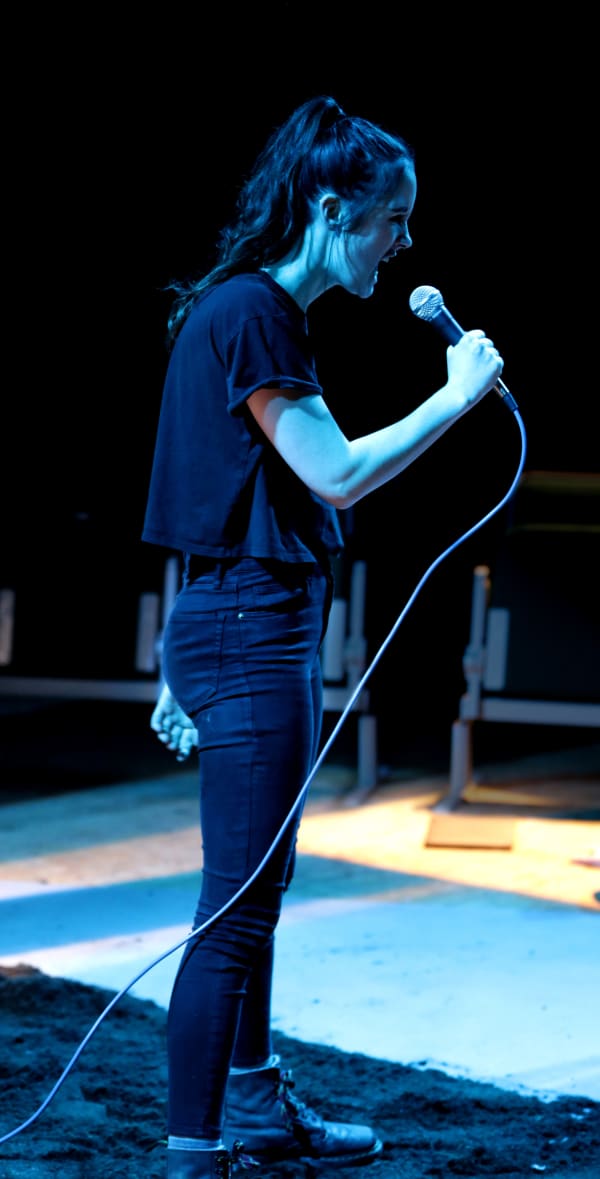 Girl in black t-shirt and jeans with a mic on stage.