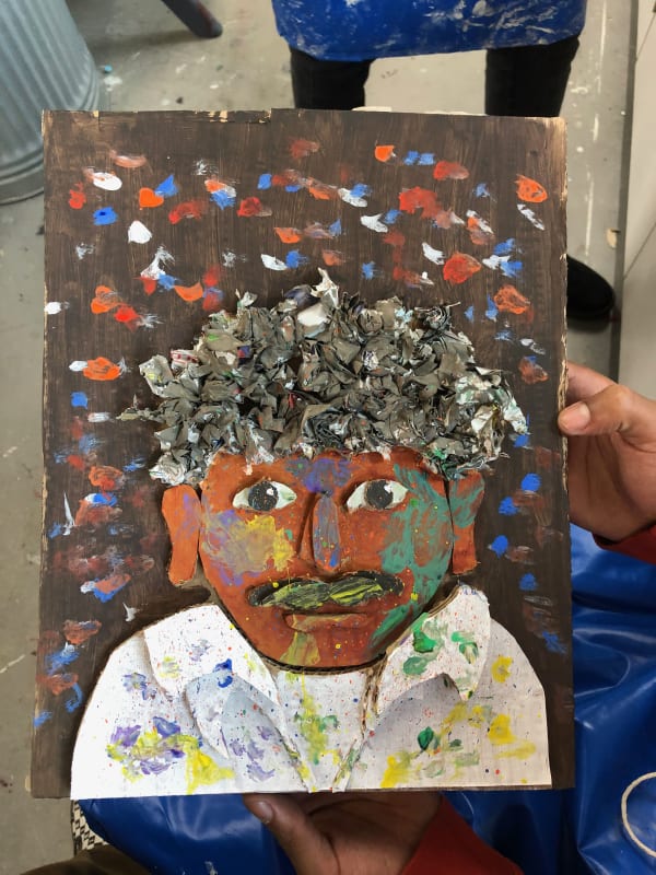 a portrait painting of a man with curly grey hair. He wears a white shirt covered in paint marks. The background is brown with multicoloured spots. 