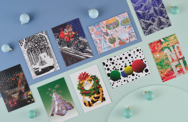 A blue and pastel green background is dotted with matching baubles. 9 Christmas cards are laid out on the table: The first card features an illustration of Santa in a tank top on a motorbike, the second is an abstract red green and yellow background with paint splatters and white letters spelling out 'NOEL', and the third is a black and white illustration of a house in a forest with a little ghost ice skating on a frozen lake, the forth is a photo of the oxford street Christmas lights through the rain, the fifth is a line drawing of Christmas dinner in lots of different colours, the sixth features four dotty colourful baubles in the Tate style and colours - red, yellow, green and blue, the seventh is a painting of a grumpy orange and black striped cat with a red bow on its head and a Christmas wreath around its neck, the eighth is a dark blue block print of mountains and the ninth is a computer generated image of a silver Christmas tree with a white to green gradient as the background.