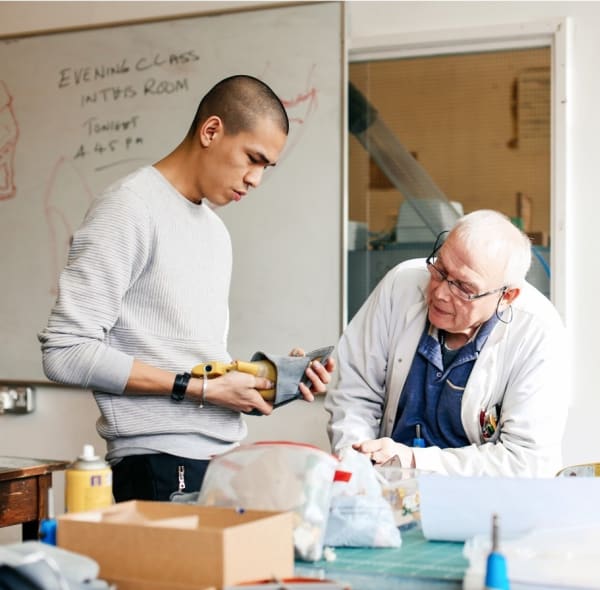 Student working on footwear in the studio with a man next to him inspecting the work.