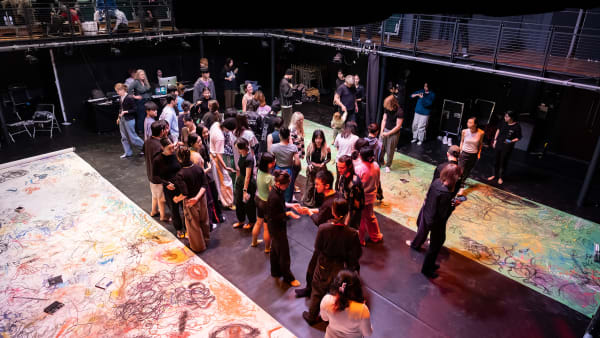 Image shows an aerial shot of a large group of people standing in a studio theatre with 2 long sheets of paper, both of which have writing and drawing on them. 