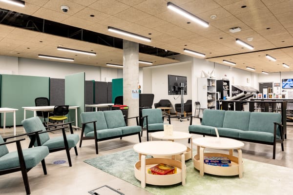 View of the workshop and study room featuring room dividers creating private desk and workstation areas. Busy students are hidden from the camera to avoid disruption. The space includes a seating area with light teal blue upholstery, bookcases and cabinets filled with student work, large workbenches, and a versatile conference screen on wheels.