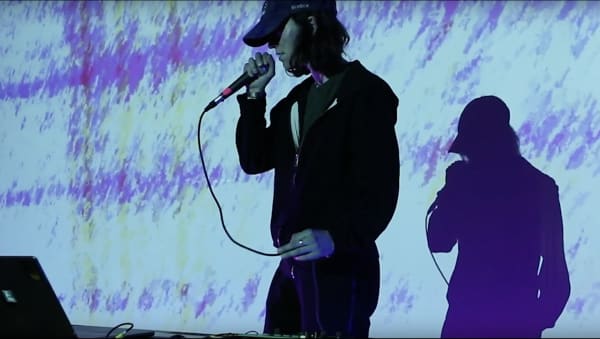 A photograph of a person standing with a microphone in front of a projector that emits a purple texture