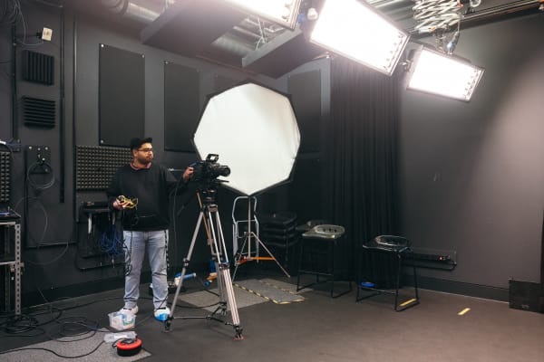 A student is setting up a professional camera and tripod. There is a large flash lighting source to their right and studio lighting on above. There are 2 tall stools to the right and cables and equipment walls also in view.