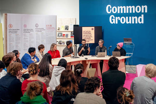 Panelists in discussion seated around a table under a sign in caps stating 'Common Ground'. An audience watches on