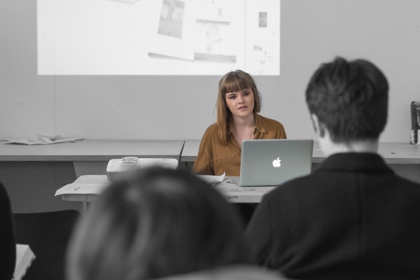 Woman seated at desk with projector and laptop, answering questions from audience