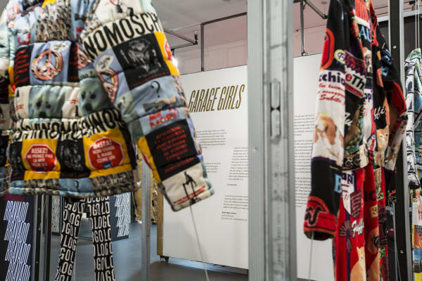 close up of Moschino graphic and text printed denim outfit and quilted down jacket. in the distance a display wall of text titled Garage Girls.