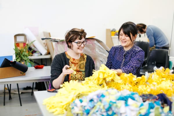 Two women sitting laughing at a table piled with yellow and blue fabric garlands