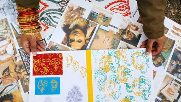 a pair of hands with gold bangles decorating the wrists, places images decorated with south asian patterns on a pile of sketchbook pages. 